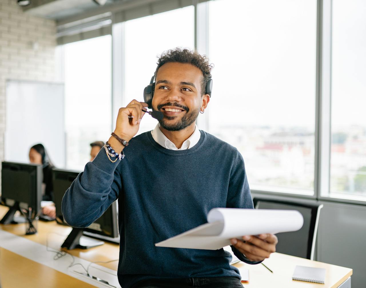 services-03 A cheerful man wearing a headset and holding a clipboard at a modern office workspace.